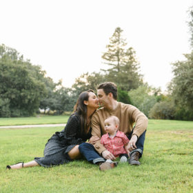 Mom and dad kiss while son looks off to the distance while sitting on grass in Sacramento
