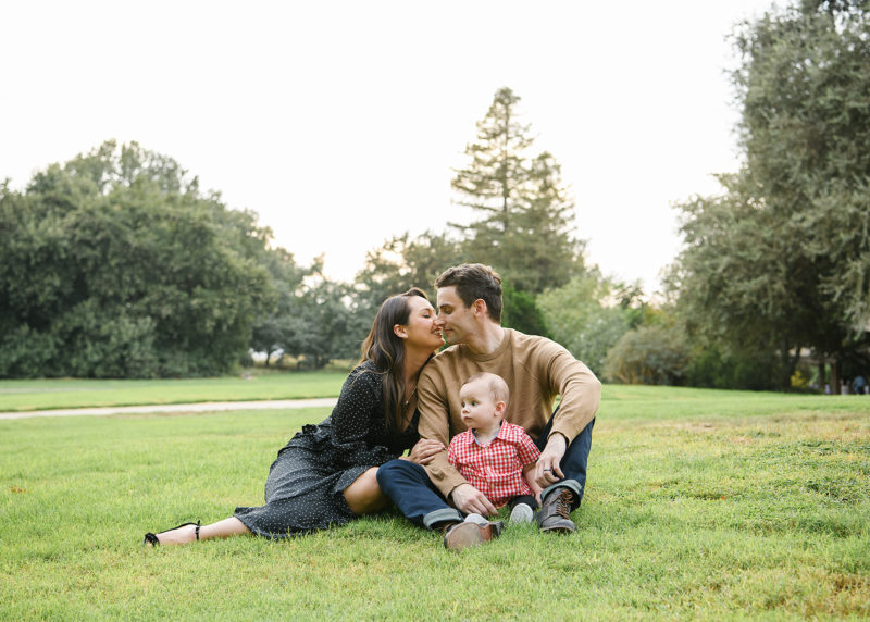 Mom and dad kiss while son looks off to the distance while sitting on grass in Sacramento