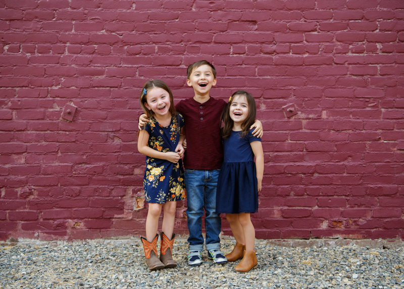 Three children laughing at the camera while standing in front of purple brick wall in midtown Sacramento