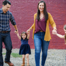 Mom and dad hold daughters’ hands walking in front of purple brick wall in midtown Sacramento