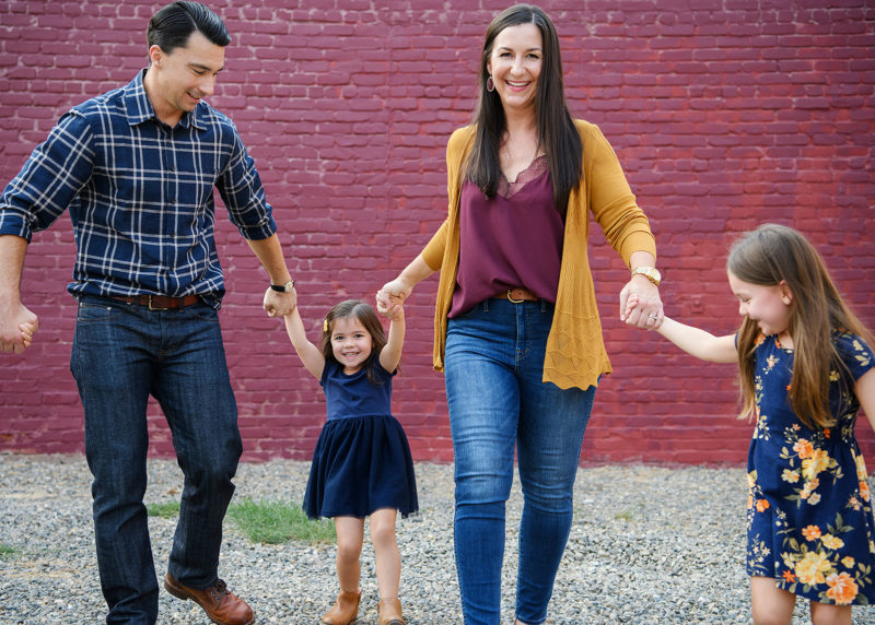 Mom and dad hold daughters' hands walking in front of purple brick wall in midtown Sacramento