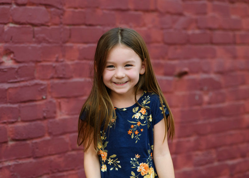 Little girl smiling directly at camera in front of purple brick wall in midtown Sacramento