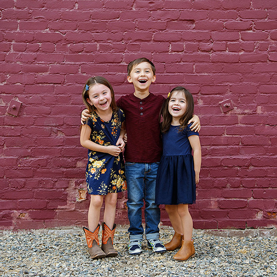 Three children laughing at the camera while standing in front of purple brick wall in midtown Sacramento