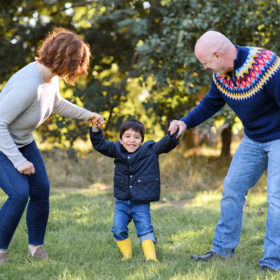 Aunt and dad hold little boy’s hand as he smiles at the camera with trees in the background in Davis Arboretum