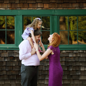 Dad holding daughter on shoulders as mom looks at her in front of green window trim and wood shingles Empire Mine State Park