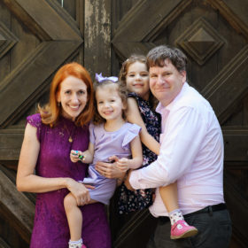 Mom and dad carry daughters as they smile for camera in front of wooden door at Empire Mine State Park