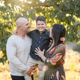 Mom and dad look at son and carry him while underneath large tree during sunset in Sacramento