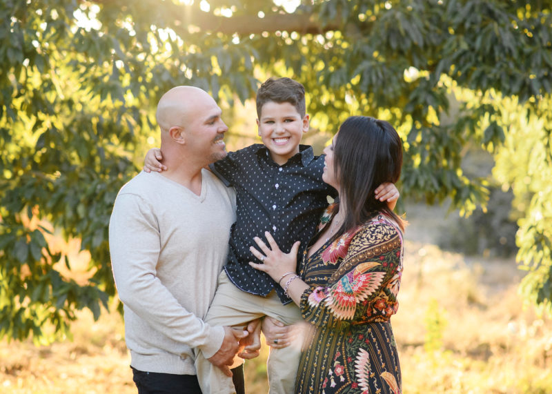 Mom and dad look at son and carry him while underneath large tree during sunset in Davis