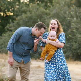 Mom laughing whole dad kisses baby boy’s hand in dry grass in Sacramento