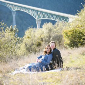 Pregnant woman and husband lying on dry grass with bridge in background Sacramento
