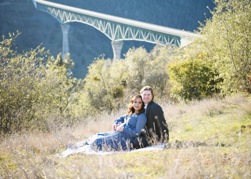 Pregnant woman and husband lying on dry grass with bridge in background Auburn