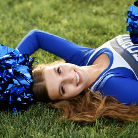 High school senior girl holding pom poms and wearing cheerleader outfit lying on grass in Sacramento