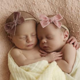 twin newborn baby girls hugging with pink bow headbands on a blanket