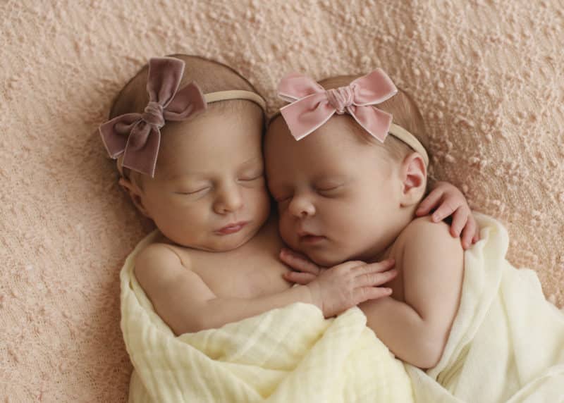 twin newborn baby girls hugging with pink bow headbands on a blanket