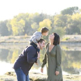 mom and son kissing on dad’s shoulders by the river
