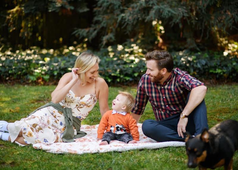 mom dad and baby boy sitting on a blanket in the grass with family dog