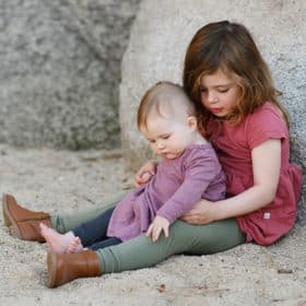 big sister and baby sister sitting on the beach looking at the sand
