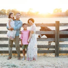 family of four posing by a wooden fence with a donkey during summer sunset