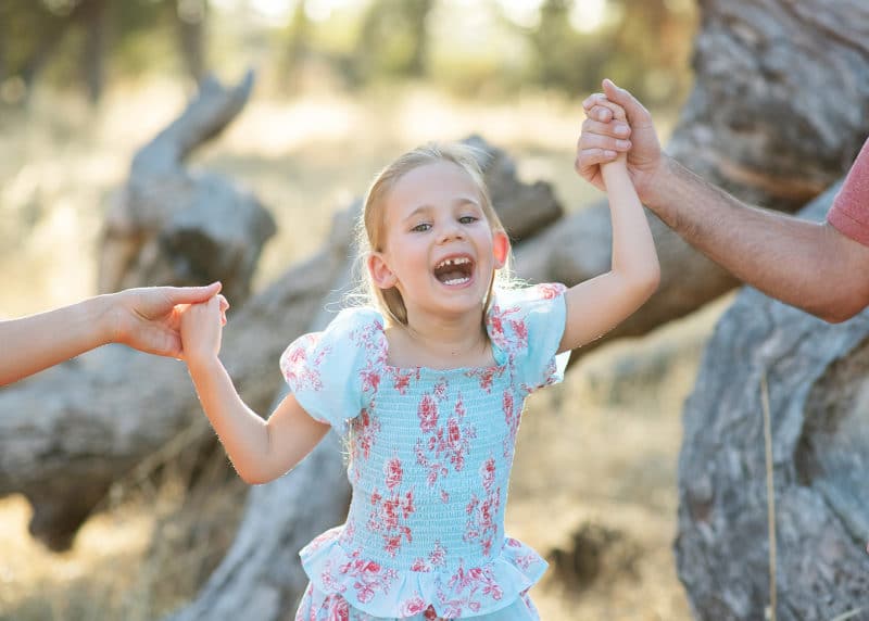 young girl laughing while holding mom and dad's hand in a field with oak logs