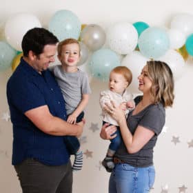 mom and dad holding two young boys in front of blue birthday balloons