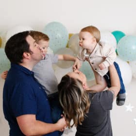 mom and dad with two young boys blowing kisses in front of birthday balloon wall