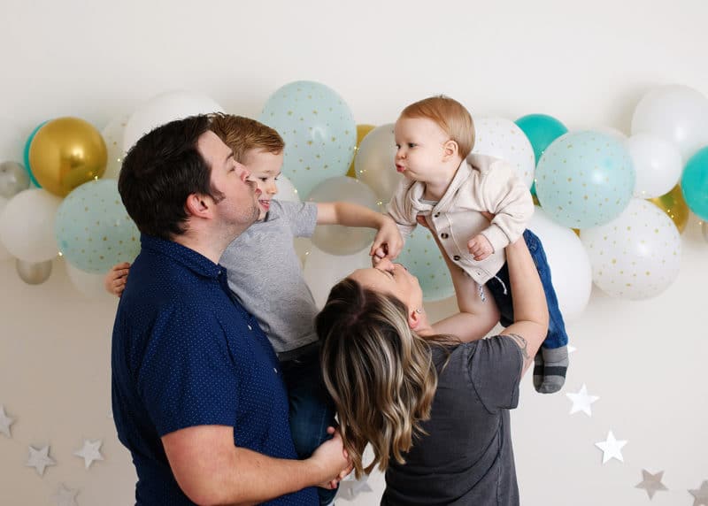 mom and dad with two young boys blowing kisses in front of birthday balloon wall