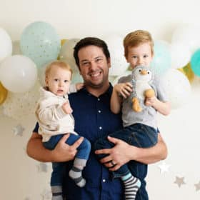 dad holding two young sons in front of blue balloons during studio photo shoot