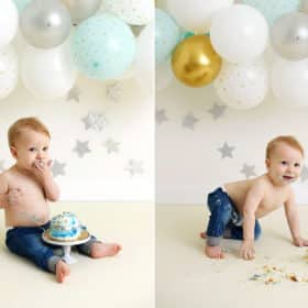 young boy licking fingers during smash cake birthday photo shoot, smiling in front of balloon garland