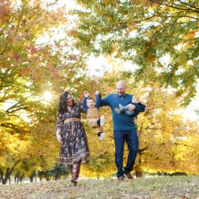 family of four holding hands swinging young son in front of fall foliage