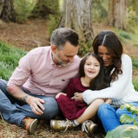 mom and dad with young girl laughing sitting on the ground in the forest