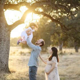 dad holding young daughter in the air with mom looking on holding pregnant belly
