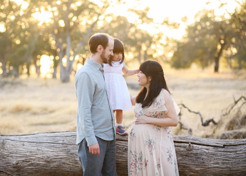 man and pregnant woman holding young daughter while standing on a log in family photo session in folsom california