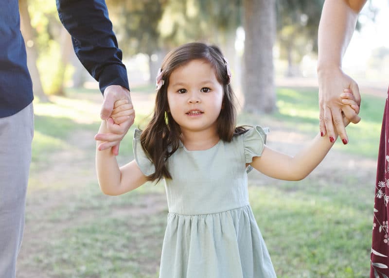 little girl holding hands with mom and dad looking at the camera during family photo shoot