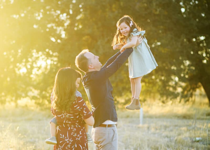 dad holding young daughter up in the air with mom and brother looking on during golden hour fall photos