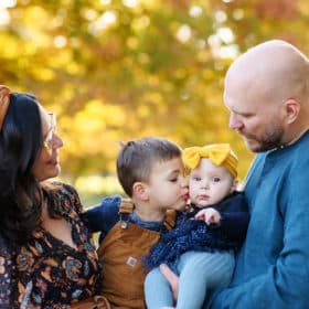 mom and dad holding son and baby daughter in front of yellow fall leaves