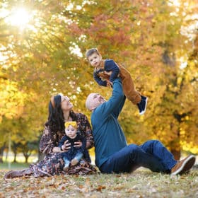 family sitting on the grass during fall photos, dad holding son in the air while mom looks on
