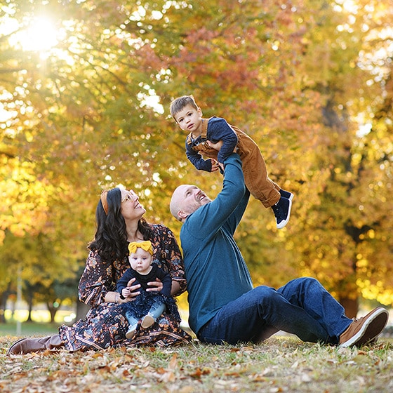 family sitting on the grass during fall photos, dad holding son in the air while mom looks on