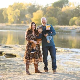 family of four standing in front of a river with fall trees during golden hour