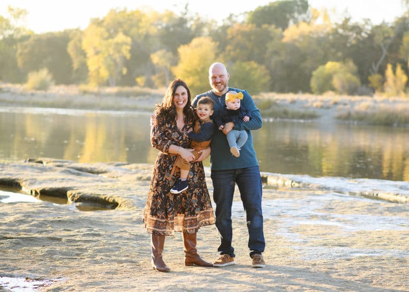 family of four standing in front of a river with fall trees during golden hour