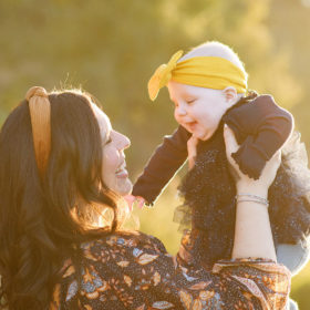 mom holding baby girl in the air and smiling, wearing yellow headbands during fall family photos