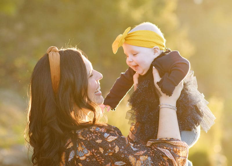 mom holding baby girl in the air and smiling, wearing yellow headbands during fall family photos