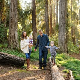 mom and dad with two young boys walking along fallen logs in the forest in davis california