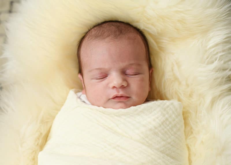 newborn baby swaddled in a blanket laying on a white fur rug during at-home newborn session