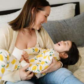mom holding young girl and laughing while sitting on bed in at-home photo session