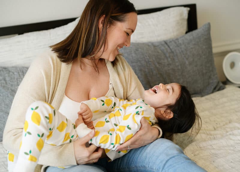 mom holding young girl and laughing while sitting on bed in at-home photo session