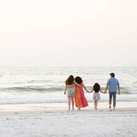 family of four standing together on the beach facing the ocean and holding hands