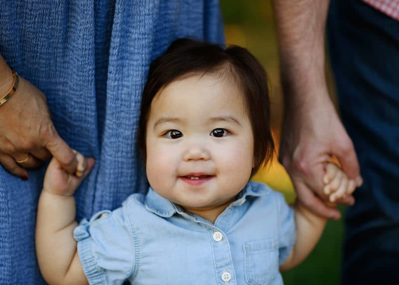 young toddler girl smiling and holding parents' hands during family photo session in sacramento
