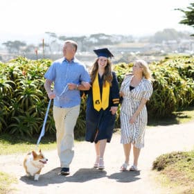 senior in college wearing graduation garb walking along the beach with parents and dog