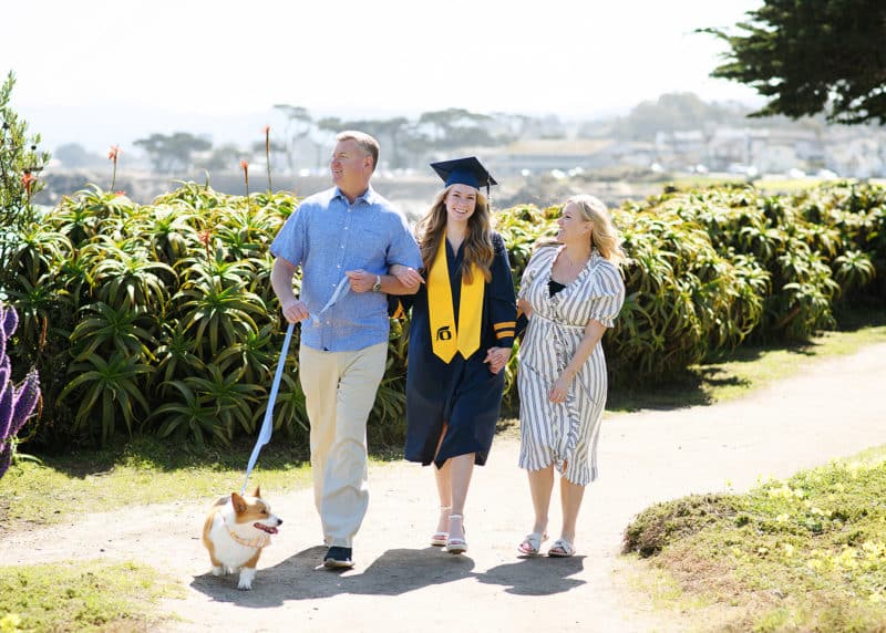 senior in high school wearing graduation garb walking along the beach with parents and dog
