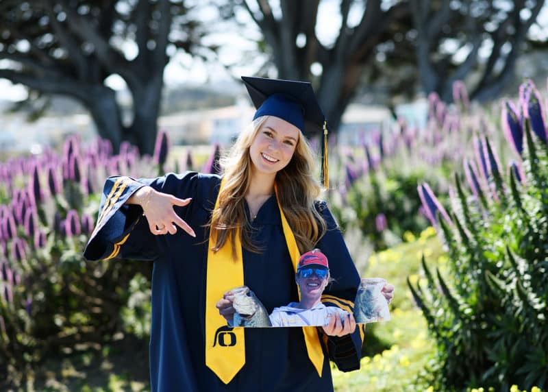senior in high school wearing graduation hat and robe and posing with picture of her brother